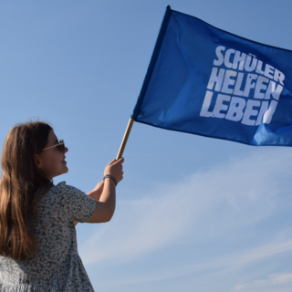 Junges Mädchen schwenkt Fahne mit Schüler Helfen Leben Text vor blauem Himmel