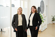 Zwei Frauen stehen in den hellen Räumen von burgenta. Eine ist blond und trägt eine Brille, rechts neben ihr steht eine brünette Frau. Beide tragen dunkle Sakkos und ein helles Unterteil.