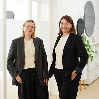 Zwei Frauen stehen in den hellen Räumen von burgenta. Eine ist blond und trägt eine Brille, rechts neben ihr steht eine brünette Frau. Beide tragen dunkle Sakkos und ein helles Unterteil.