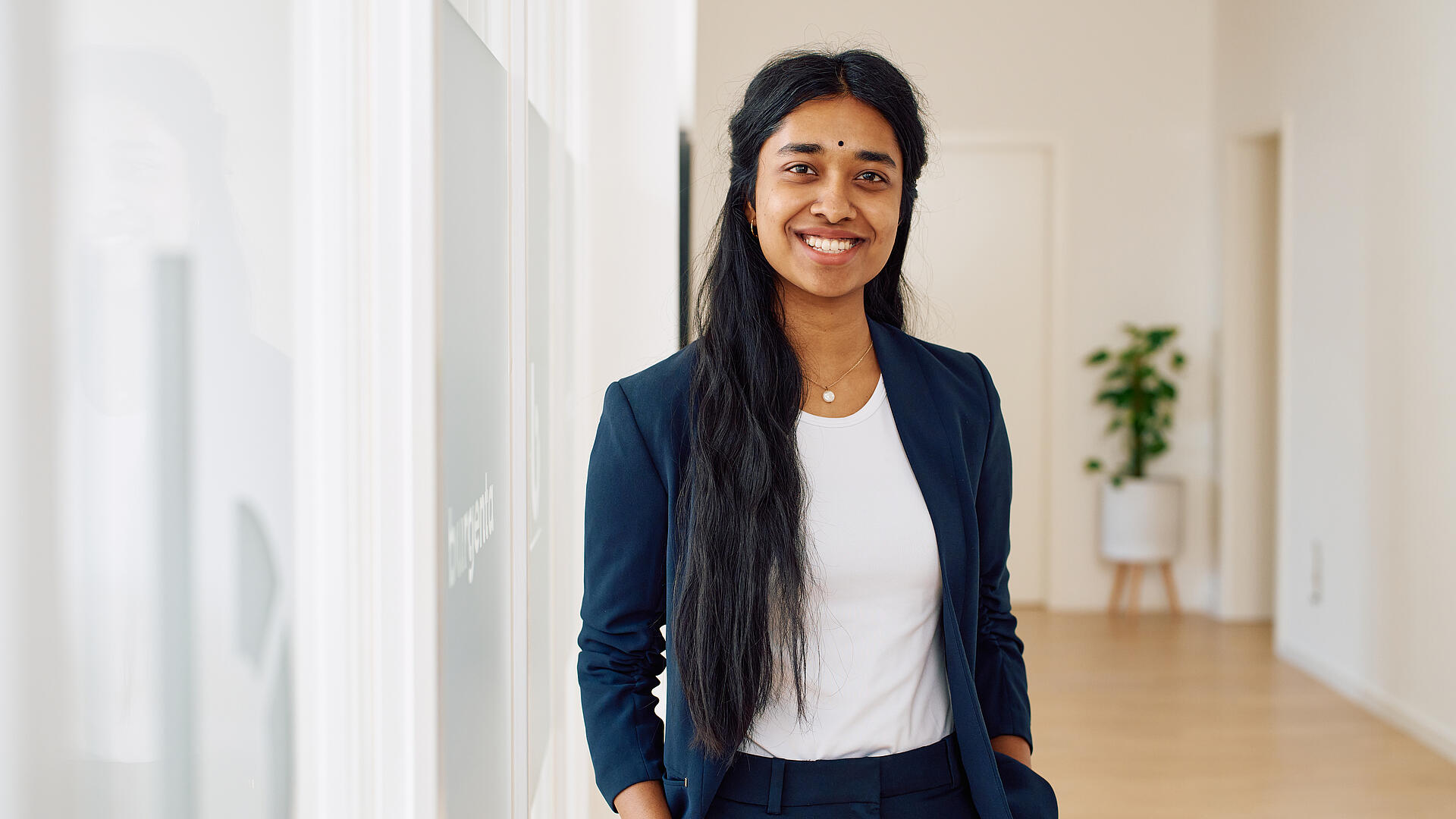 Asha steht in den Büroräumen von burgenta. Sie hat lange, schwarze Haare und trägt ein weißes Oberteil sowie einen dunkelblauen Blazer.