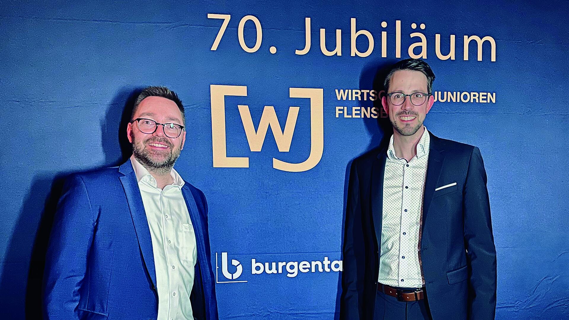 Philip Hofeditz und Jan Keirat von burgenta stehen vor einer blauen Wand. Auf der Wand ist in goldenen Buchstaben zu lesen: Siebzigstes Jubiläum Wirtschaftsjunioren Flensburg.