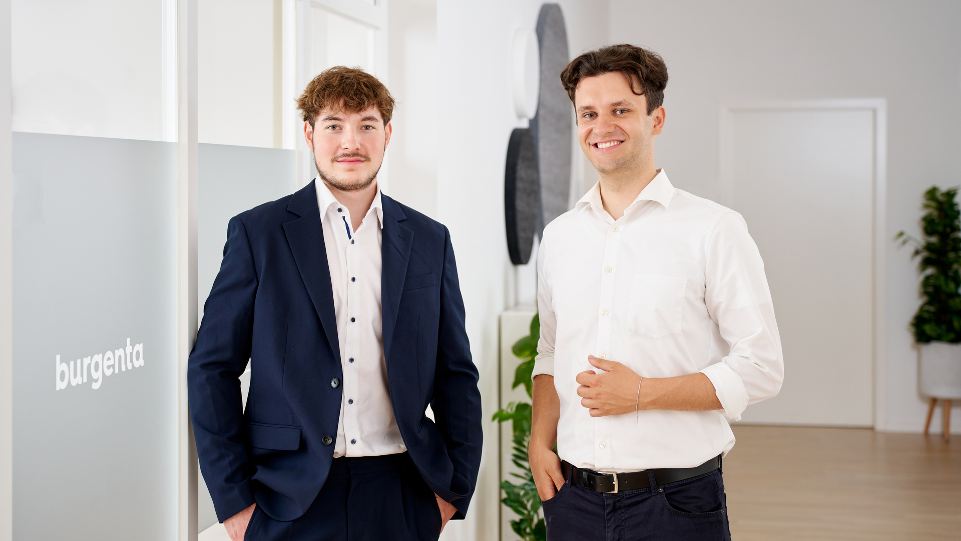 2 junge Männer stehen in Business Outfit nebeneinander, sie lächeln, im Hintergrund ist ein modernes Büro Ambiente und eine Scheibe mit Inschrift "burgenta" zu sehen