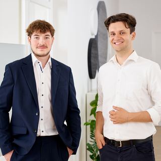 2 junge Männer stehen in Business Outfit nebeneinander, sie lächeln, im Hintergrund ist ein modernes Büro Ambiente und eine Scheibe mit Inschrift "burgenta" zu sehen