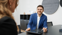 burgentas neuer Partner Till sitzt im Anzug an einem Schreibtisch, lächelt einer Person gegenüber zu und hält einen Stift sowie ein Tablet in der Hand in einem Büro von burgenta.